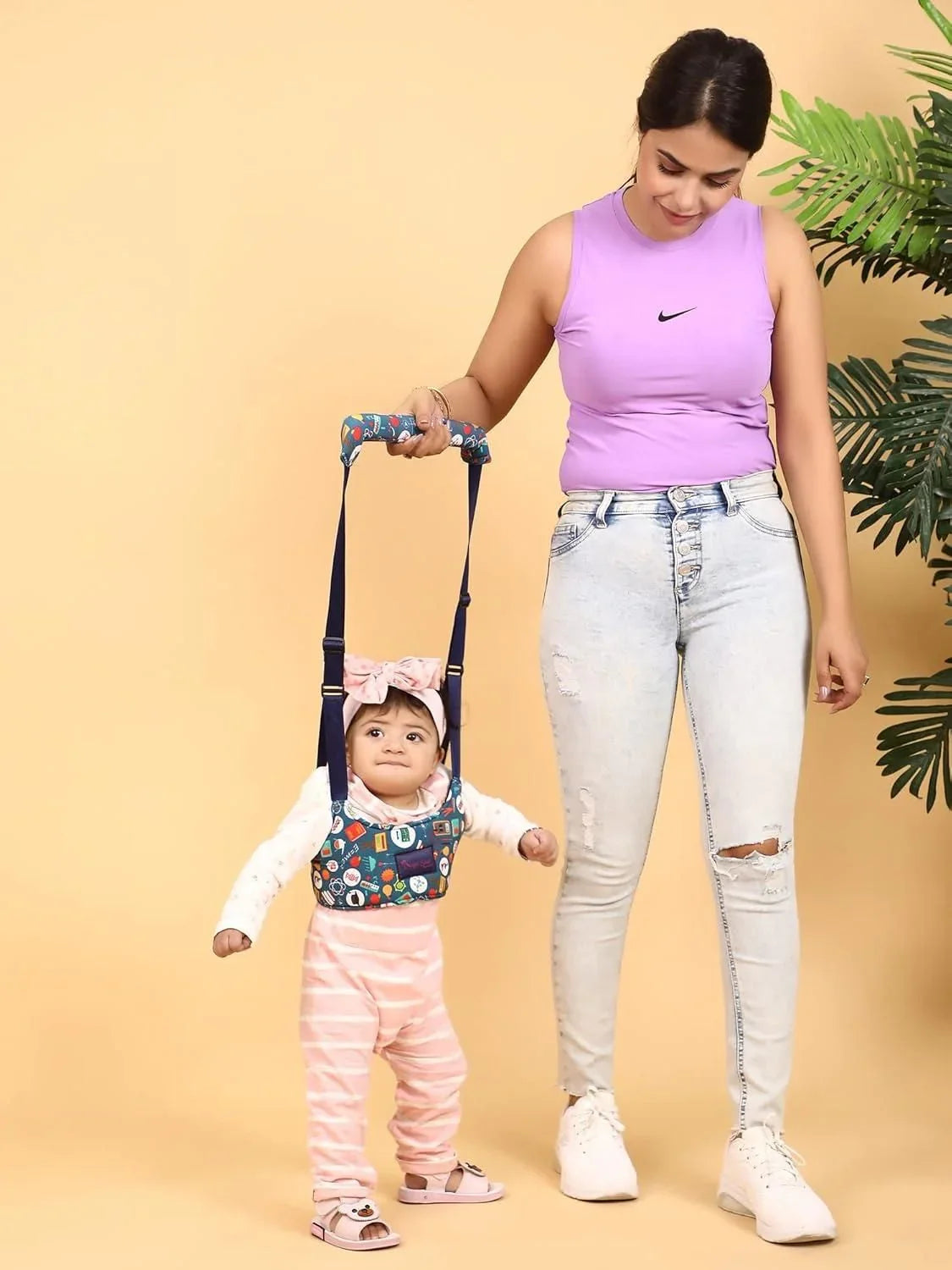 Mother in purple top helping baby girl in walking harness with beige background and plant